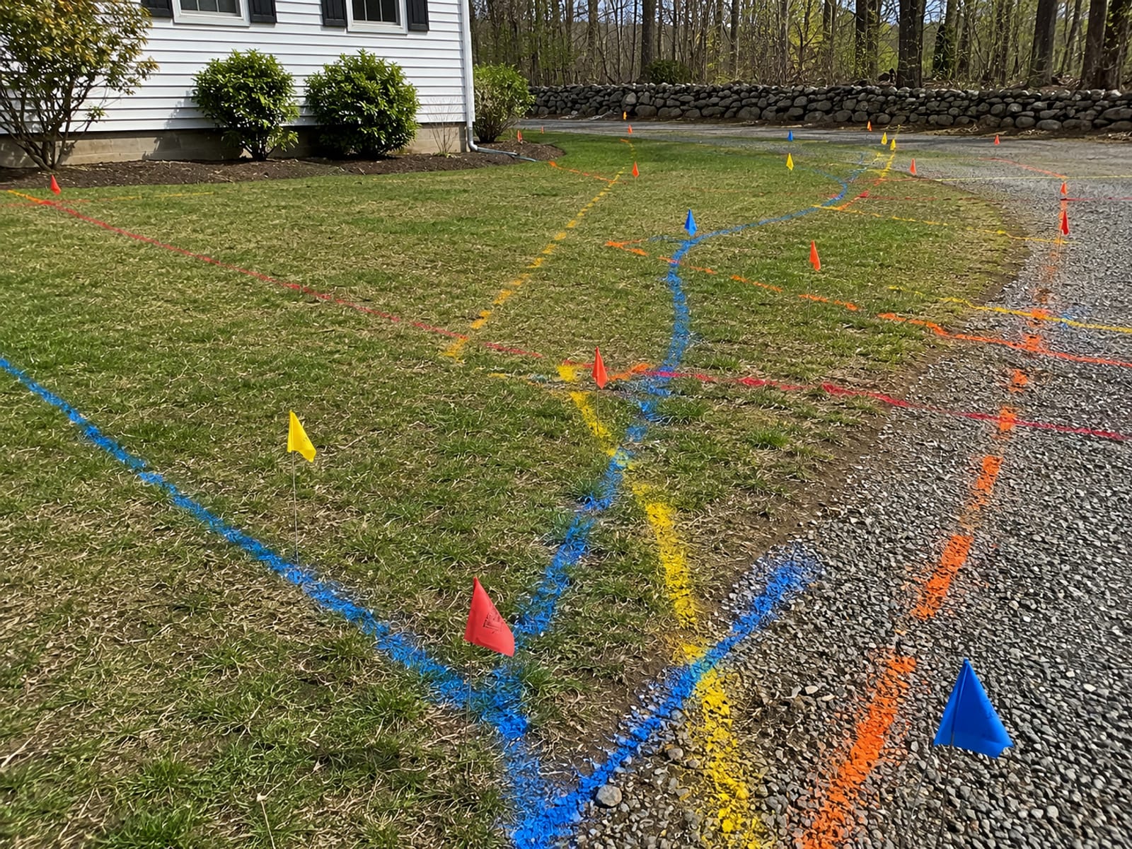 Residential yard with spray-painted utility locates in multiple colors on the grass and small colored marking flags indicating underground utility lines before excavation