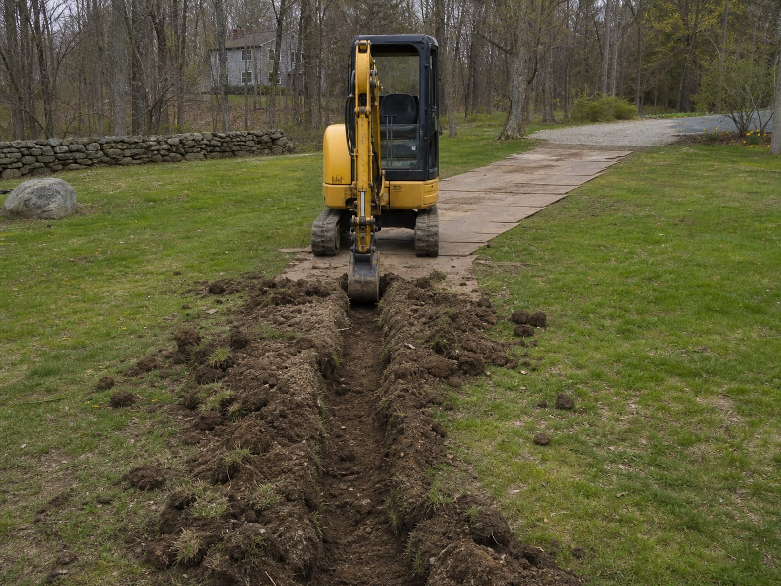 Compact excavator parked and idle at the open end of a shallow drainage trench across a rural lawn, bucket resting on the ground, excavated soil mounded along the cut