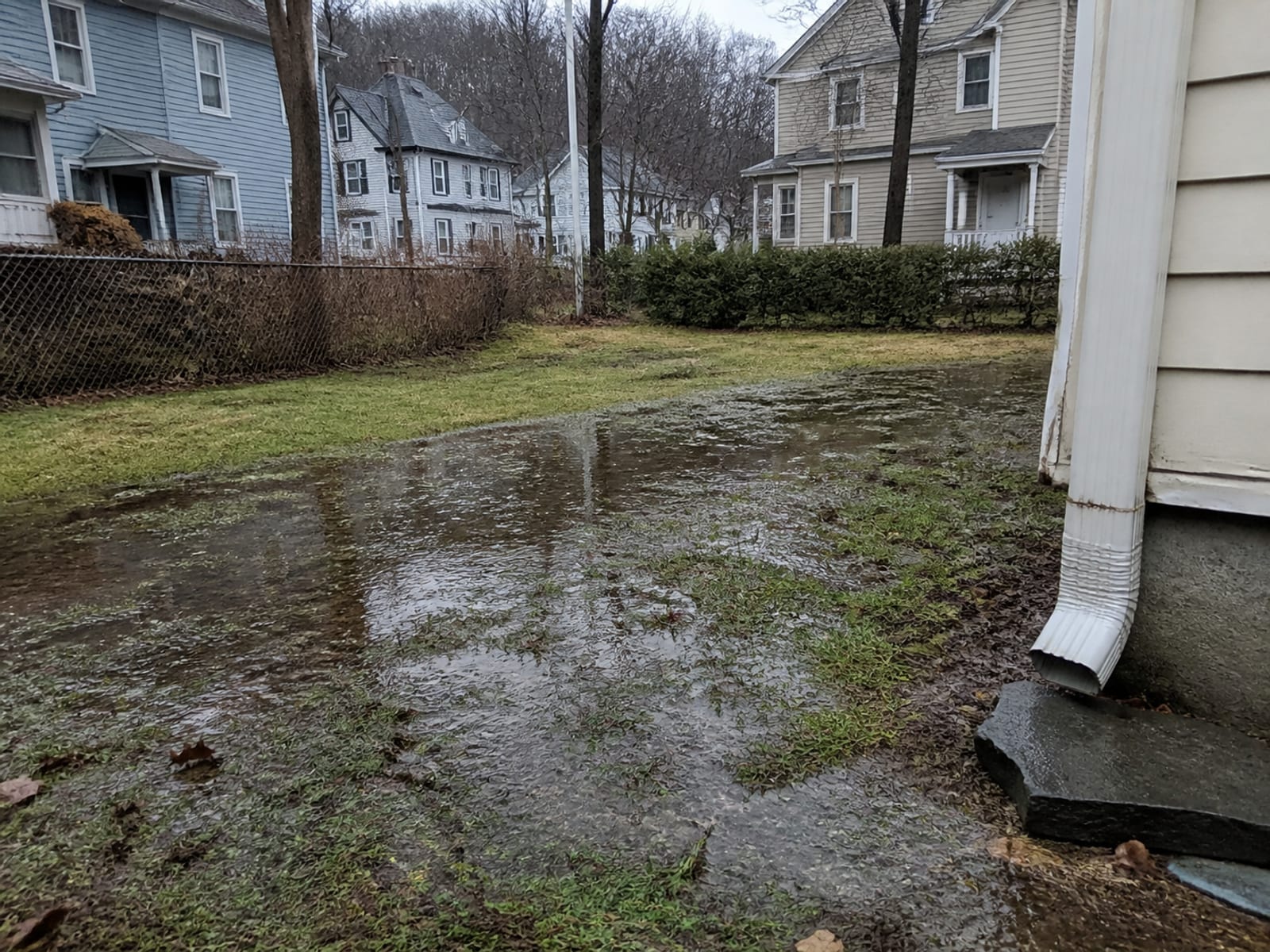 Standing water pooled across a low section of a residential back yard after heavy rain, with saturated turf and a downspout discharging nearby