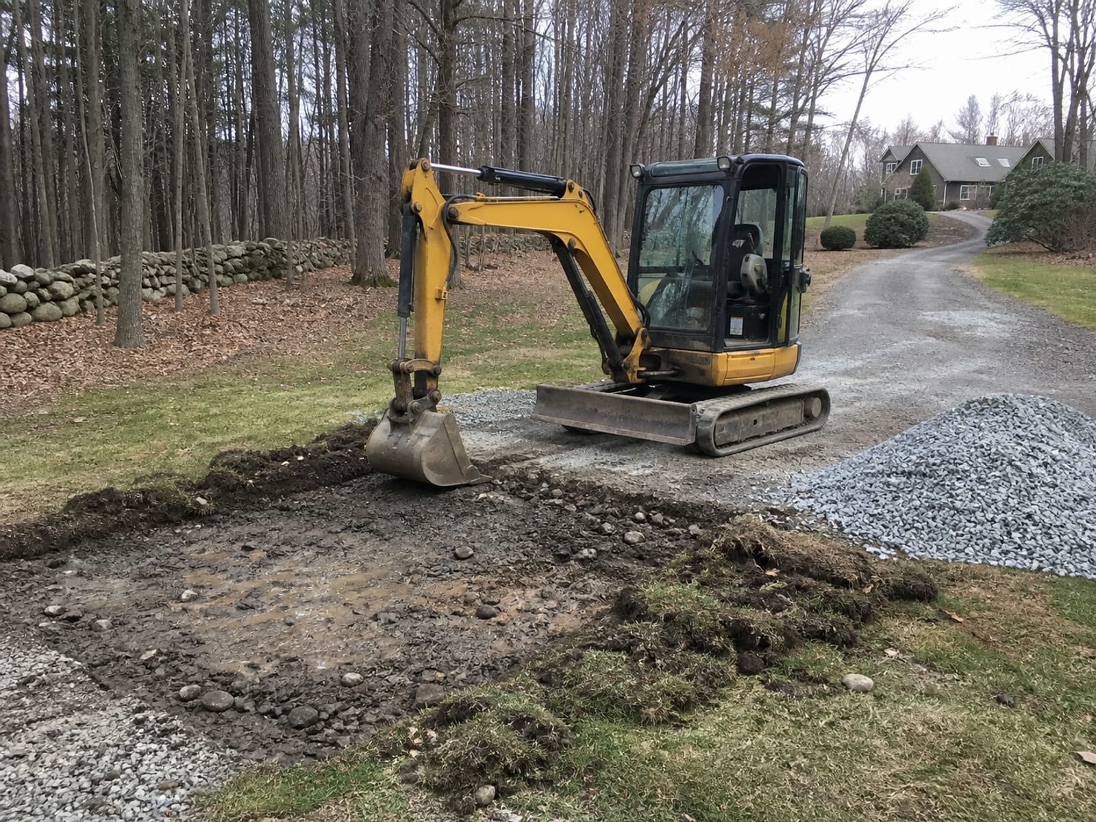 Excavator lifting out saturated stone and soil from a failed section of a gravel driveway, with clean replacement aggregate stockpiled alongside