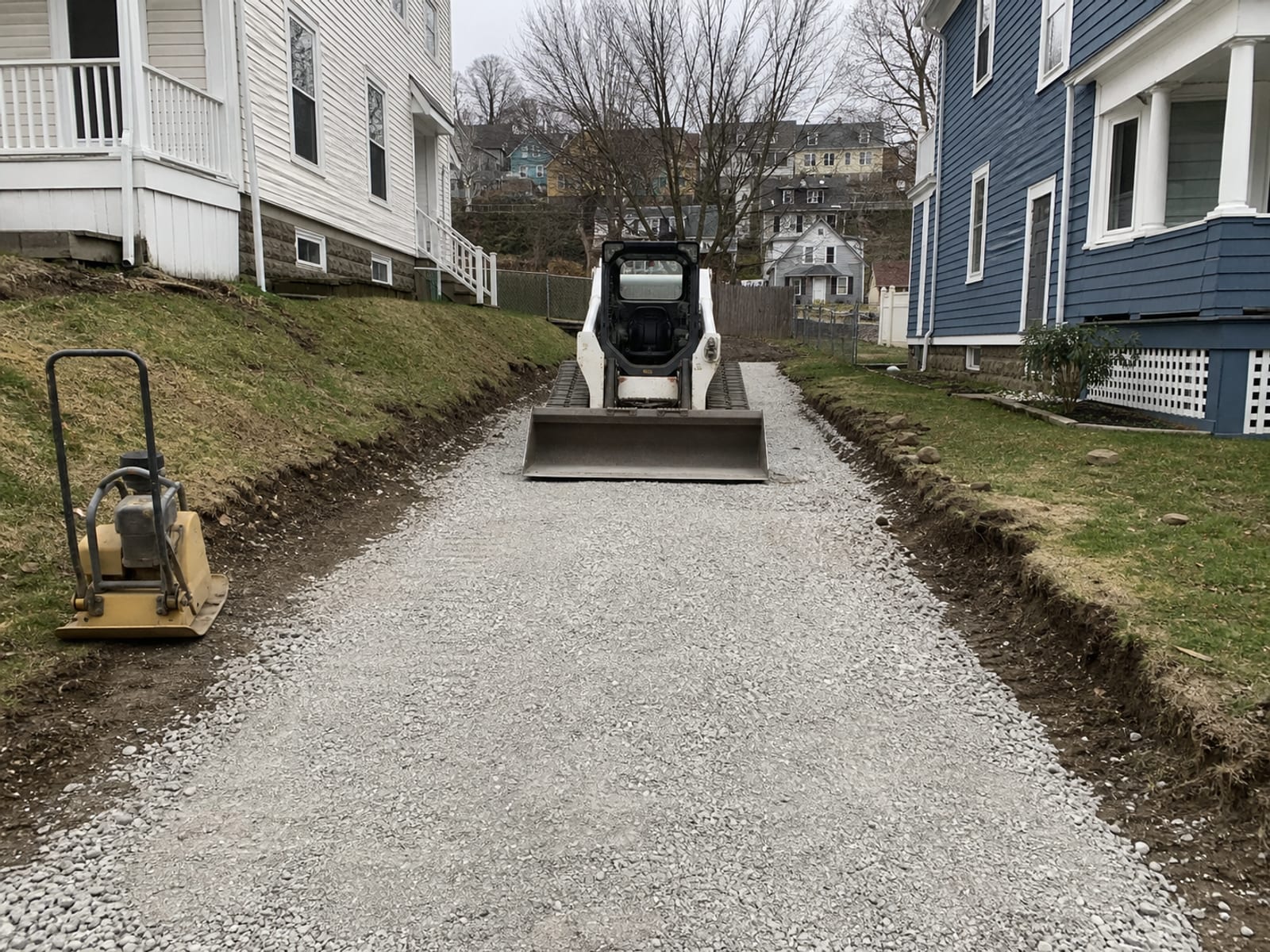 Compacted aggregate base placed along a residential driveway with a plate compactor and a compact loader parked at the open end of the run