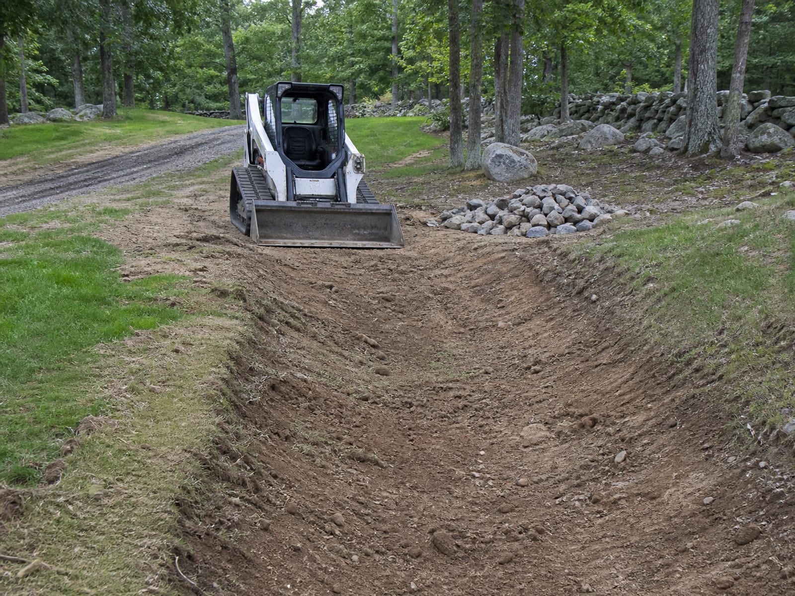 Compact track loader parked at the end of a newly shaped diversion swale above an eroded slope, bucket lowered to the ground, stone staged ready for the outlet