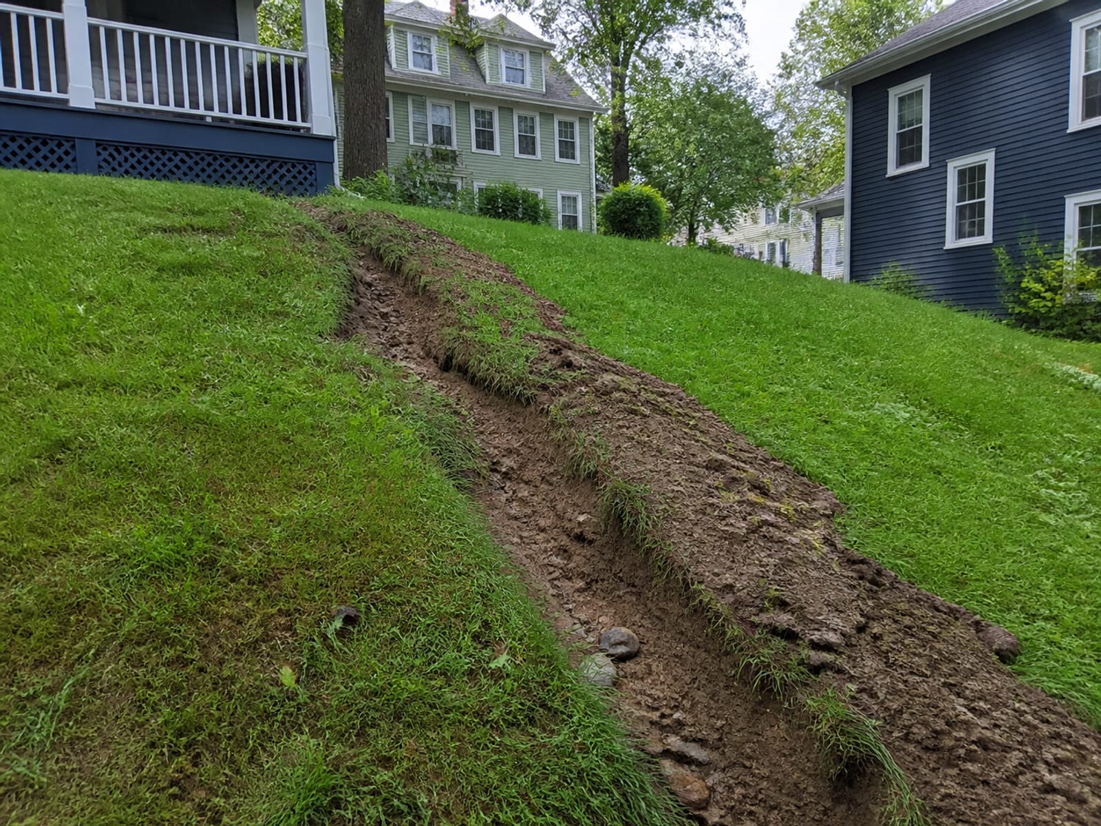 Fresh erosion channel cut across a residential slope after heavy rain, with exposed roots and loose soil carried down toward a lawn edge