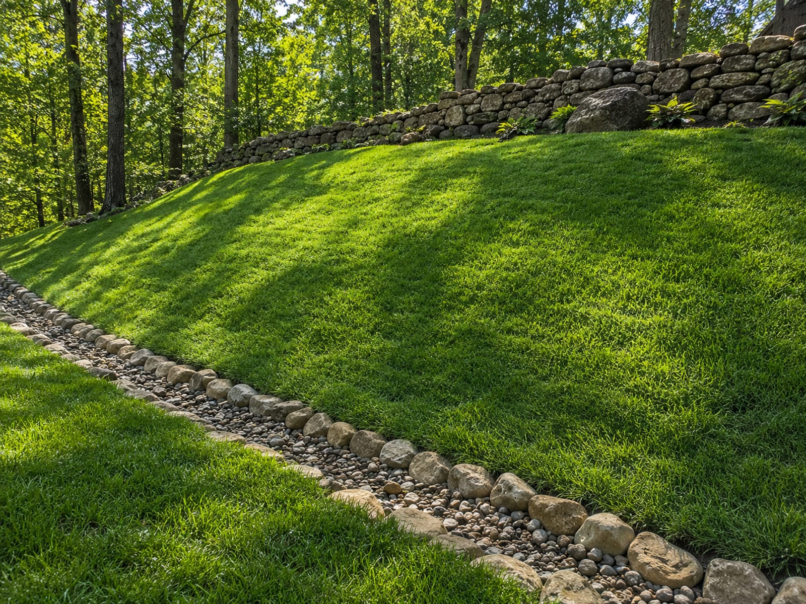 Fully stabilized residential slope in Oxford CT with dense established grass cover, stone-edged drainage swale, and mature deciduous trees framing the background