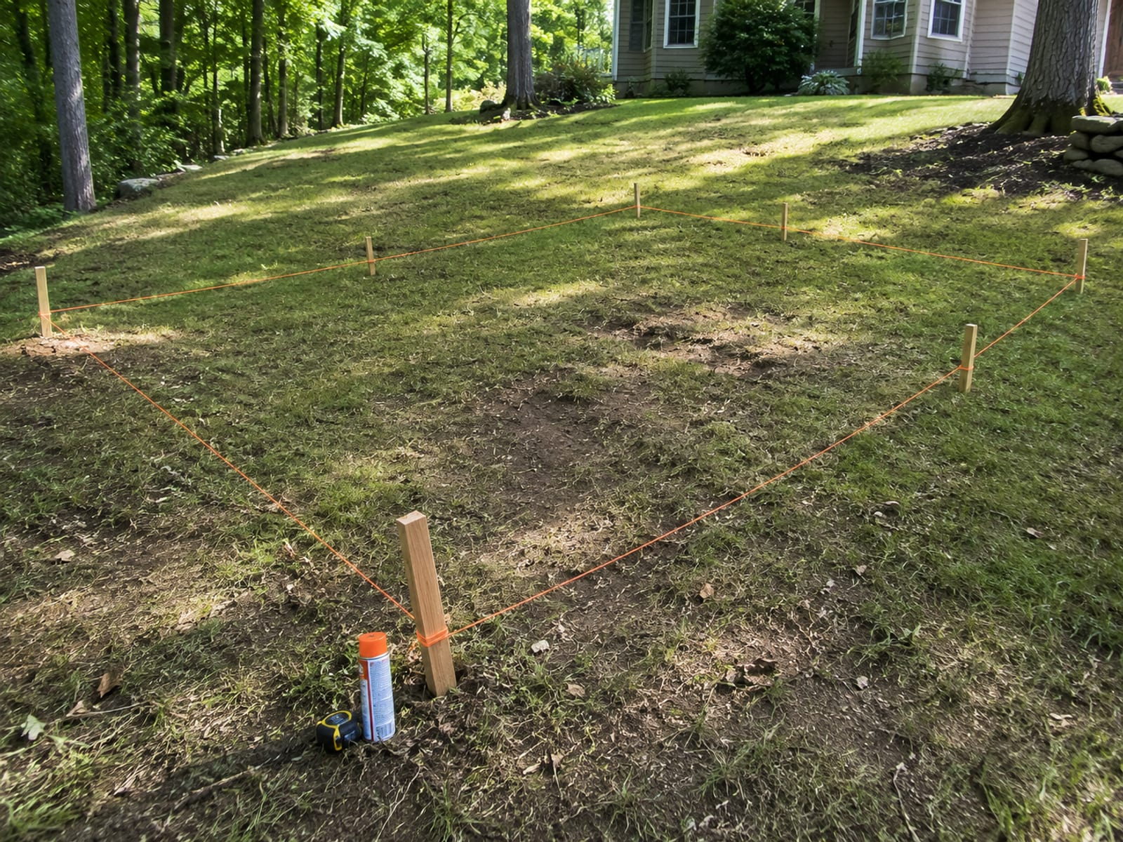 Rectangular patio footprint marked with wooden stakes and orange string lines in a sloped Connecticut backyard before excavation begins