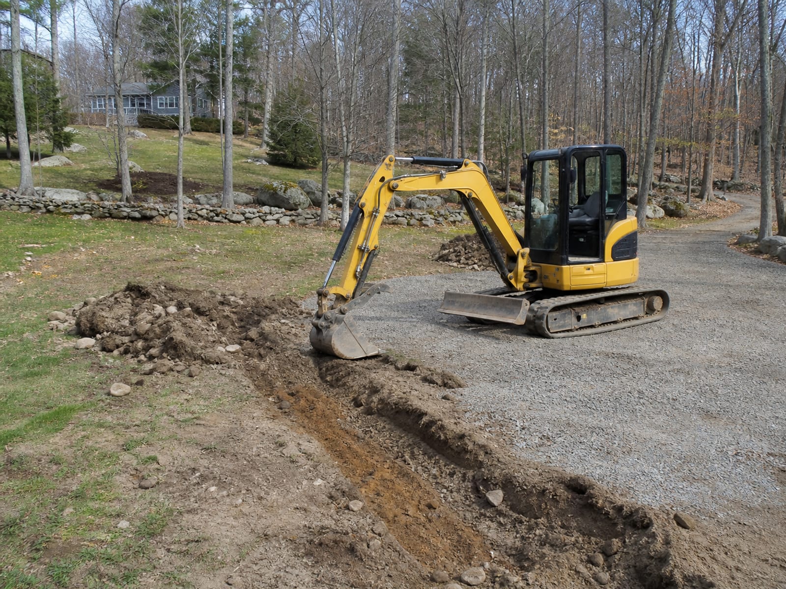 Tracked excavator parked beside an open excavation on a sloping wooded lot, boom lowered and bucket resting on the ground, excavated material mounded well back from the edge