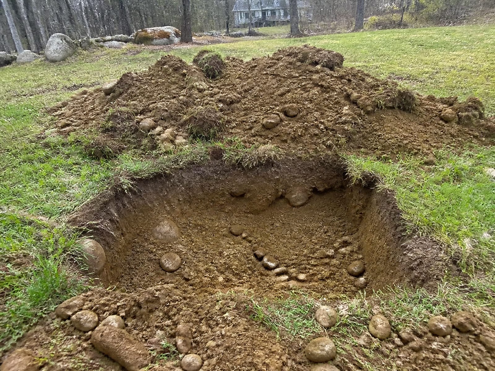 Open test pit on a wooded rural lot showing stony subsoil in the excavated face, with the removed material set back from the edge