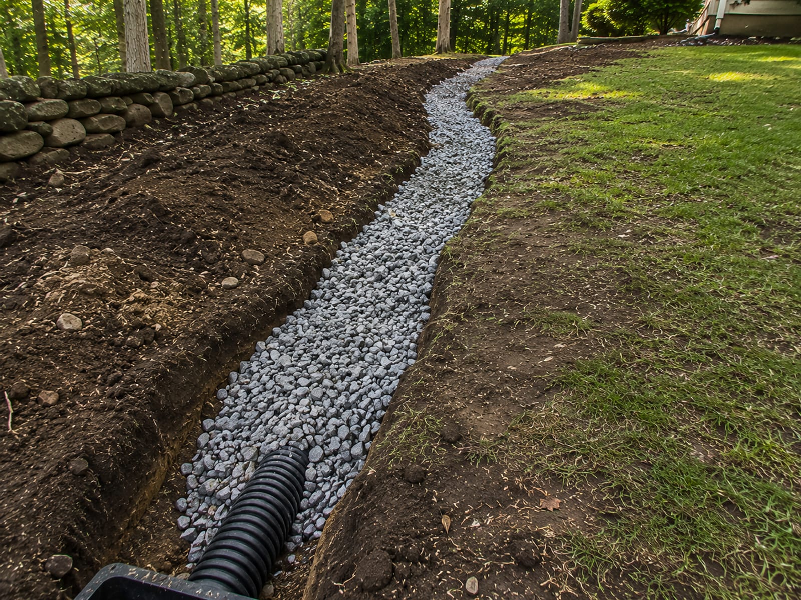 Freshly installed French drain trench with perforated pipe and clean crushed stone fill running along the base of a sloped residential yard in Woodbury CT