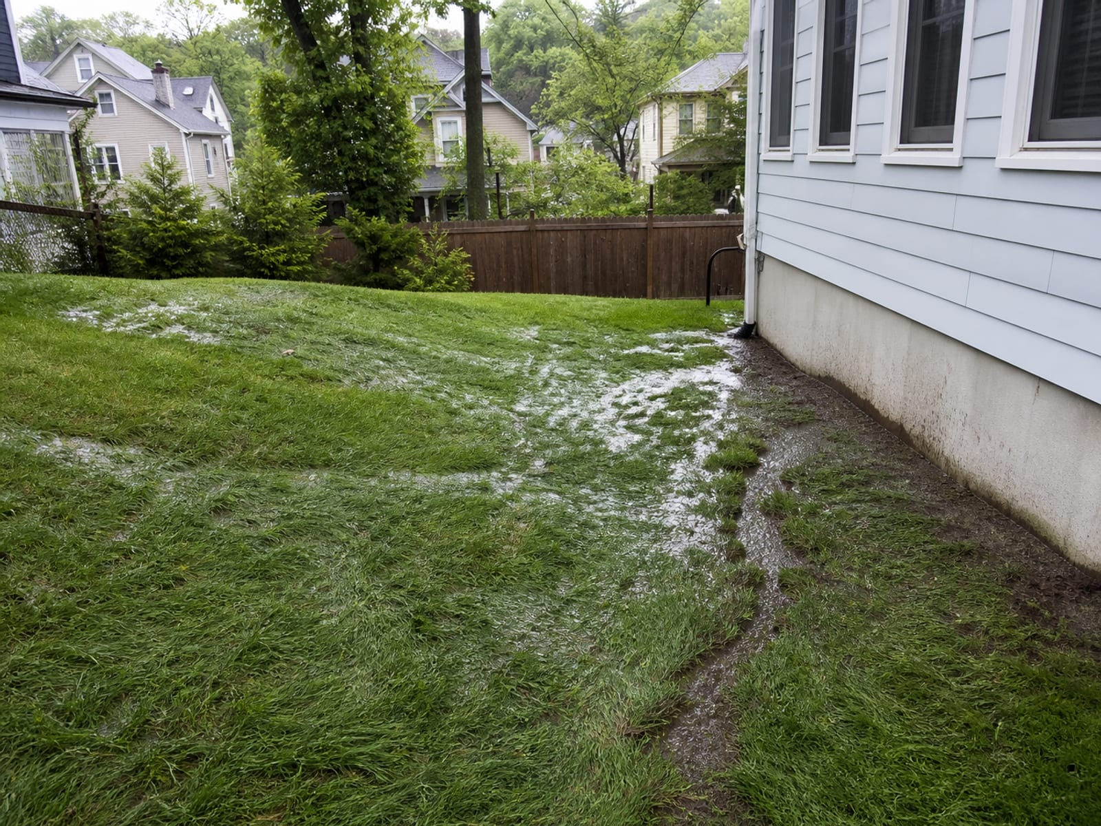 Rainwater sheeting across a residential lawn toward a house foundation, with a shallow erosion channel cut into saturated turf near the wall