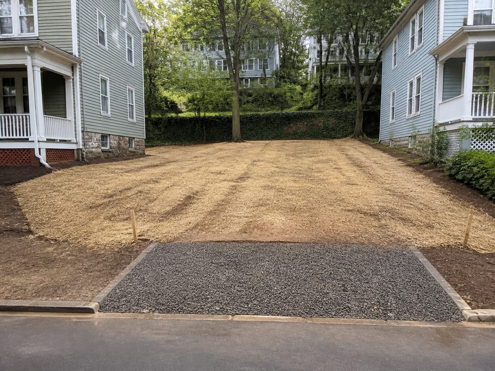 Cleared and rough graded urban parcel with property corners visible, straw over seeded soil, and a stabilized entrance at the street edge