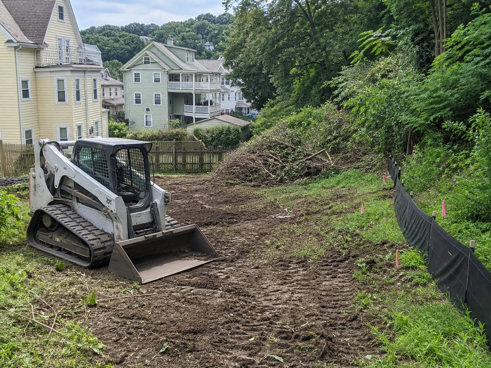 Compact track loader clearing brush inside a flagged limit on an urban parcel, with silt fence along the downhill boundary and cut material stacked for hauling