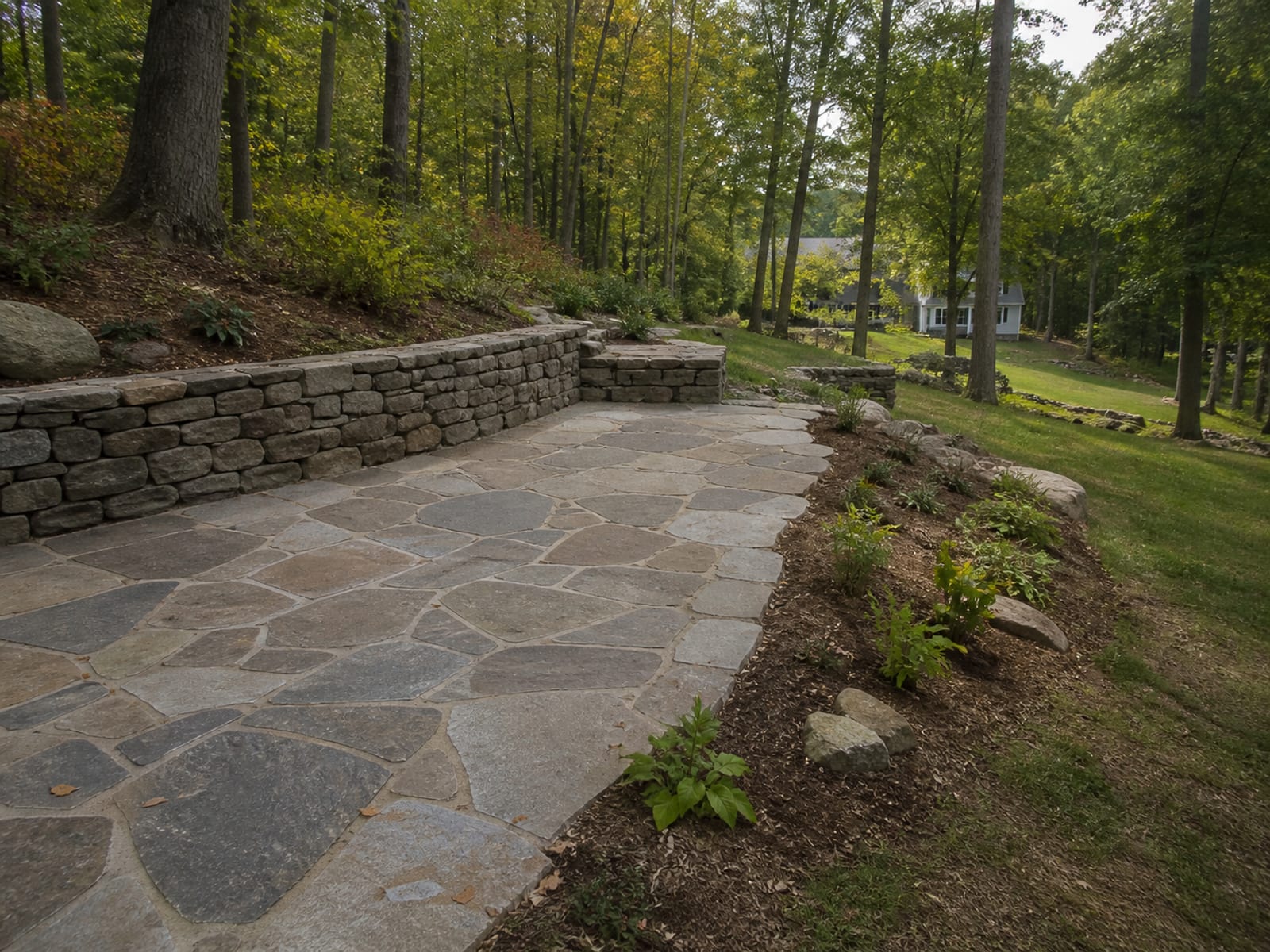 Completed stone patio set into a wooded slope with a low retaining wall along the cut bank and planted ground falling away on the downhill side