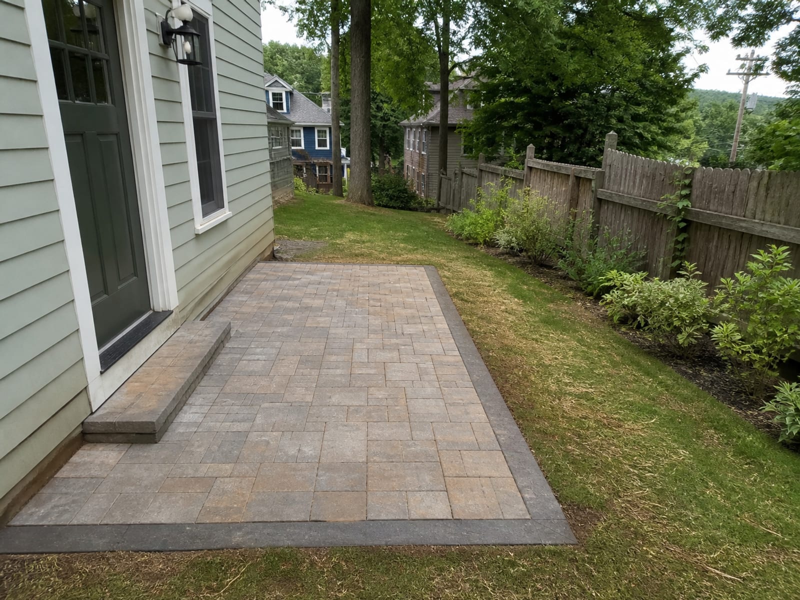 Completed paver patio outside a back door on a small city lot, set below the threshold and pitched away from the house toward a planted edge