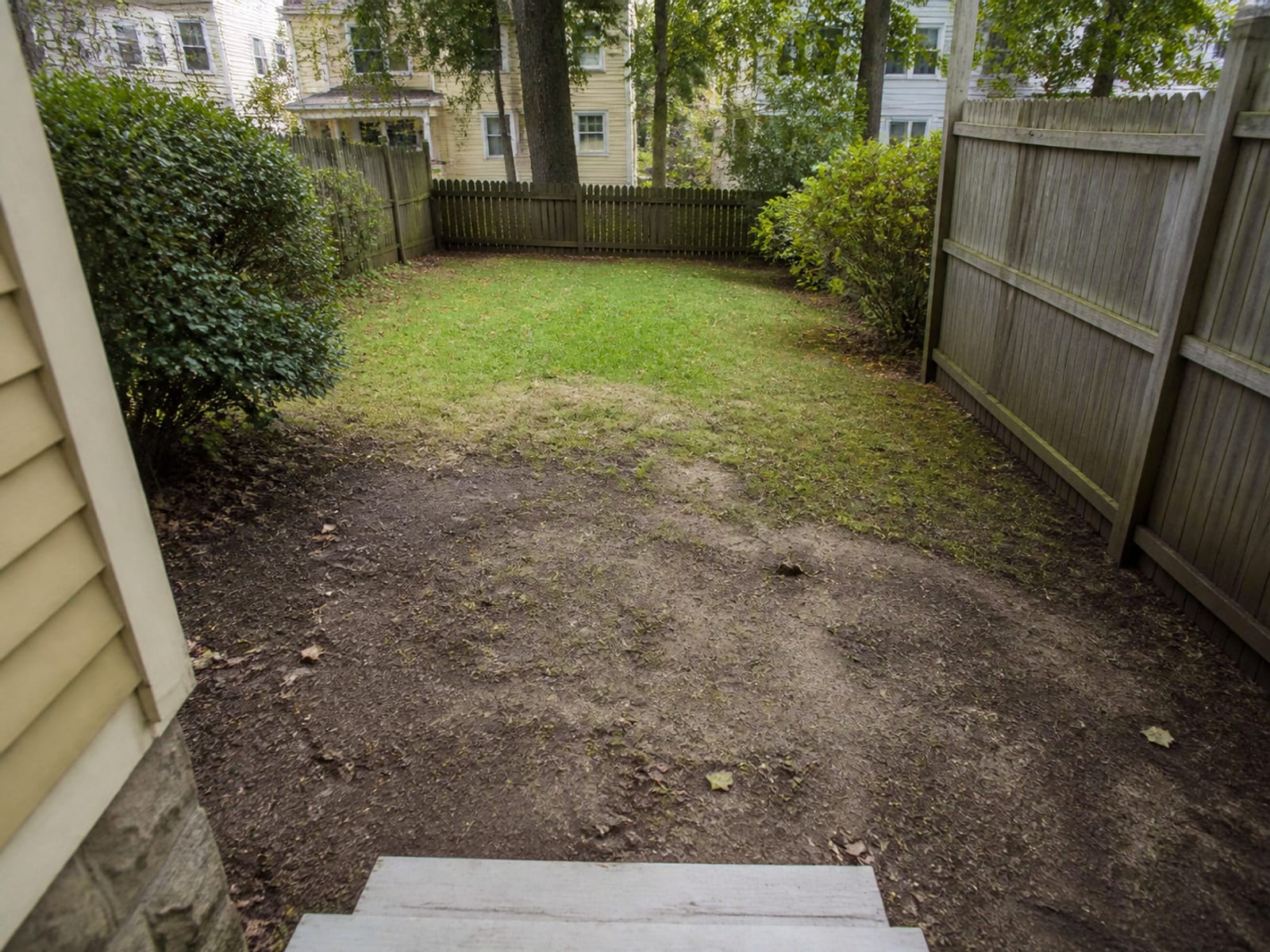 Small urban back yard sloping toward the rear of a house, with a single step down from the door and worn muddy ground where a patio is planned