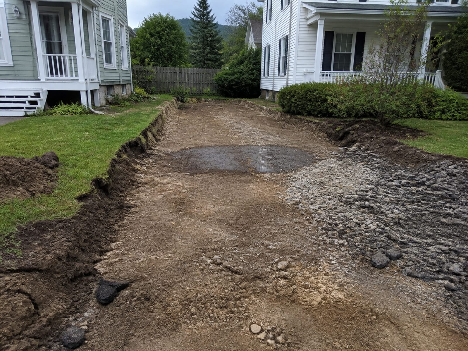 Driveway excavation with the old asphalt removed, showing the exposed subgrade with a soft damp area and old fill material at one edge
