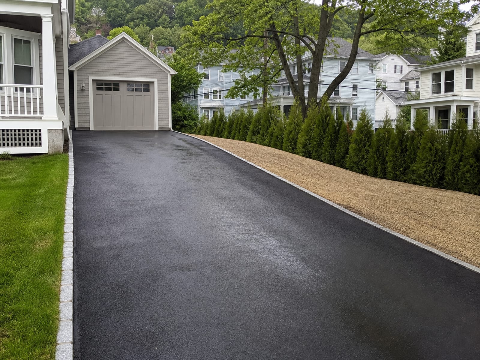 Newly paved asphalt driveway on a compact urban lot, sloped away from the garage with a clear edge and water shedding to the side