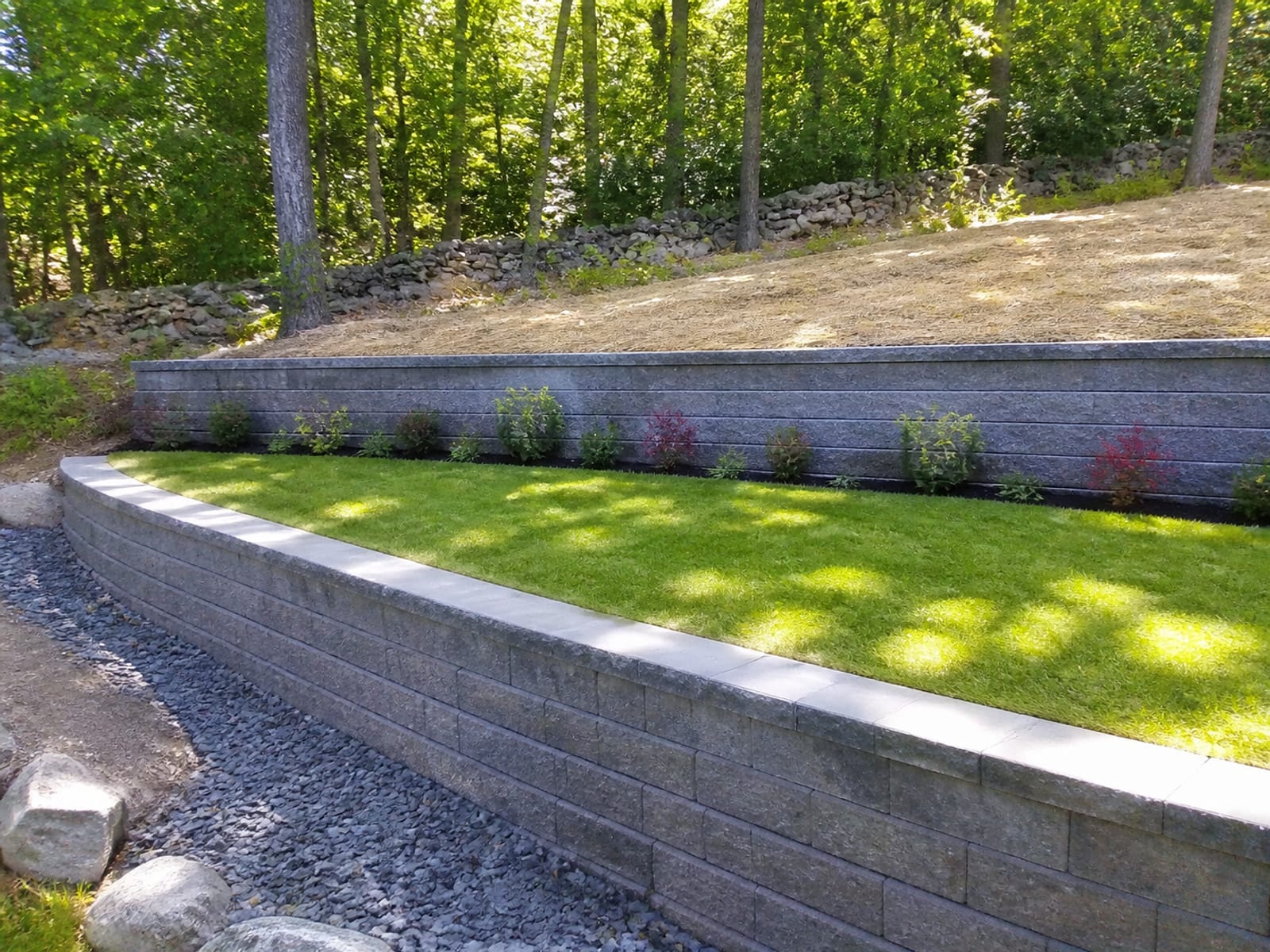 Two stepped retaining walls on a wooded slope with a planted level area between them and seeded ground above the upper wall