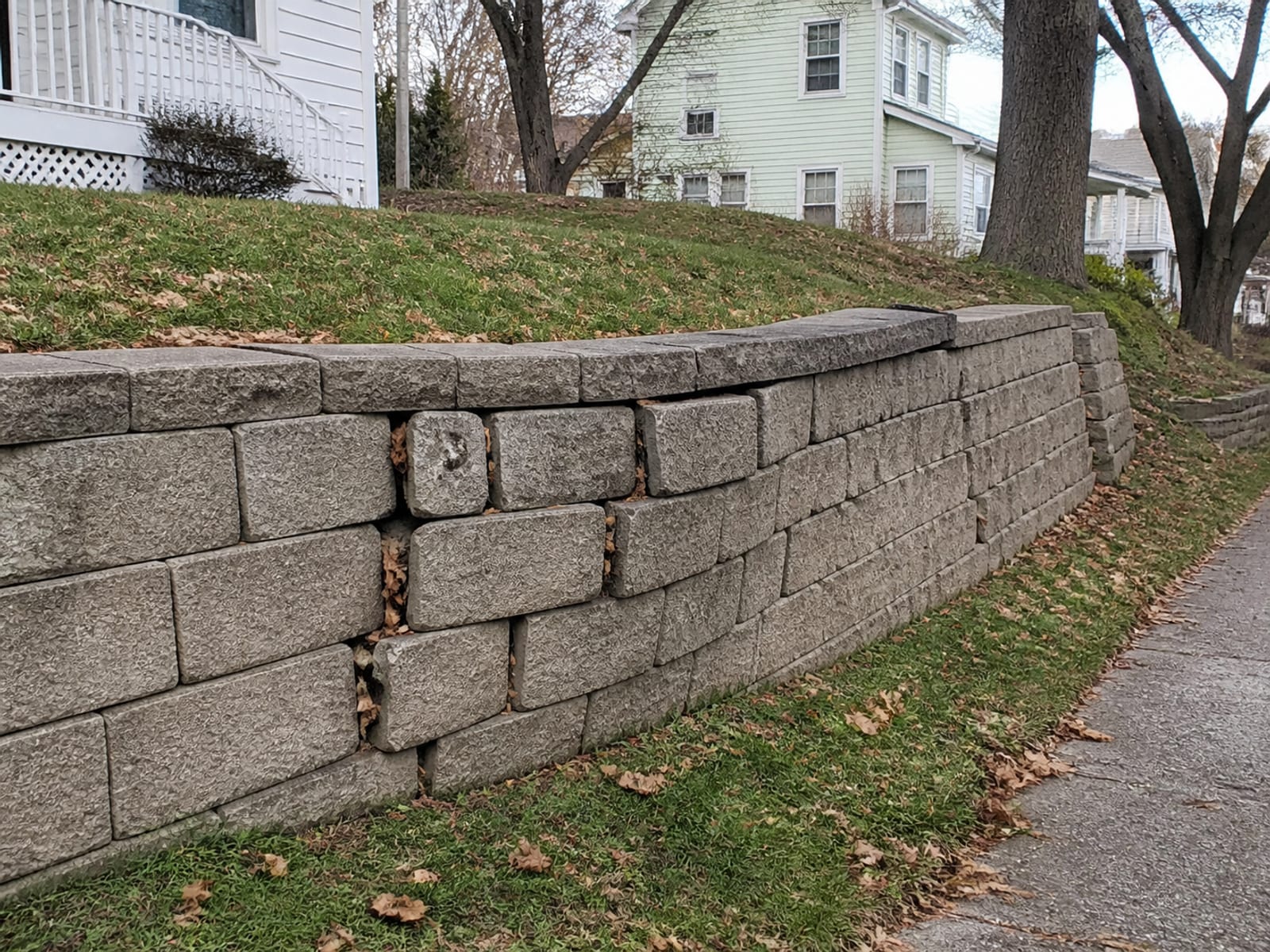 Older residential retaining wall showing forward lean and a bulge in the middle courses, with soil visible through open joints and displaced stone at the top