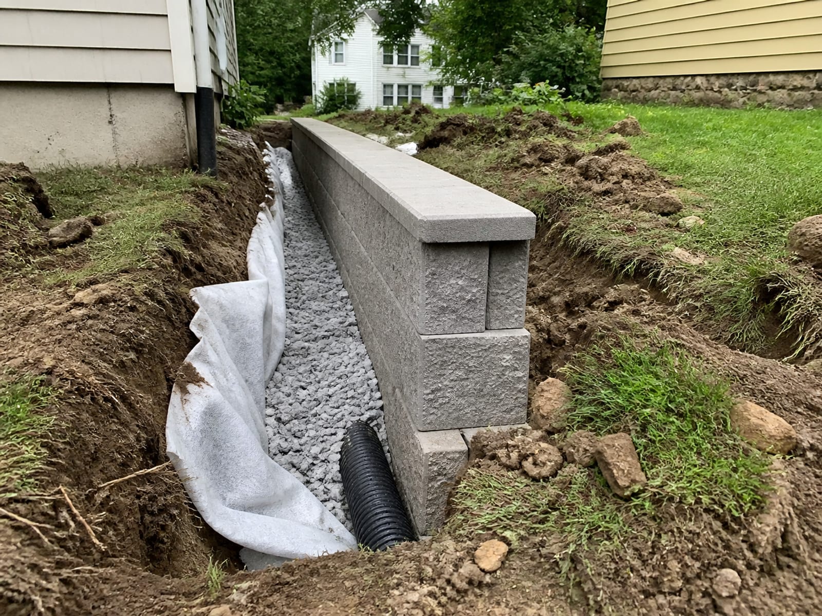 Cutaway view of retaining wall construction showing clean drainage stone, perforated collector pipe, and filter fabric placed behind the wall units