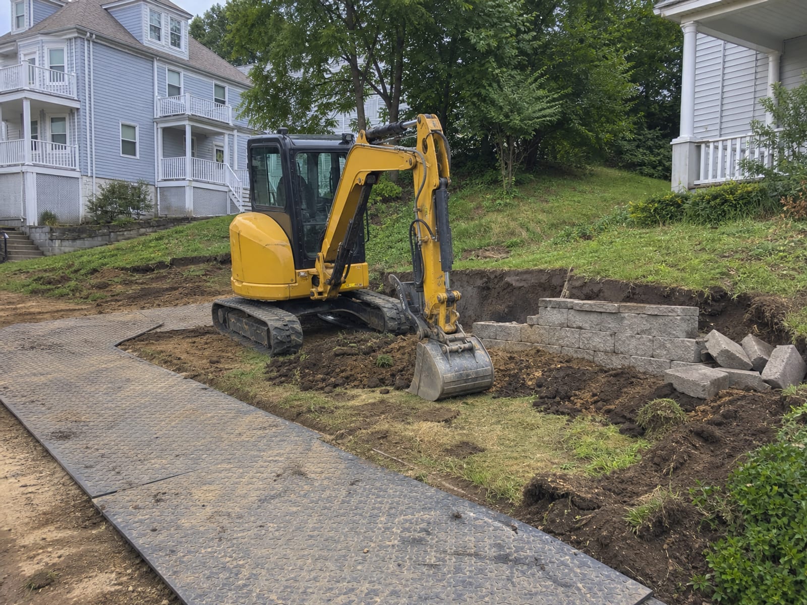Compact tracked excavator parked beside a partly demolished retaining wall on a residential slope, bucket resting on the ground beside removed block and exposed subgrade