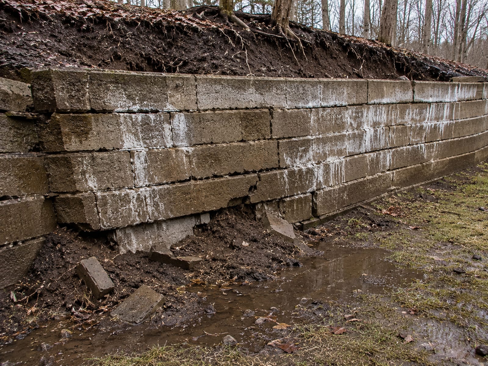 A deteriorating concrete block retaining wall visibly tilting forward with cracked sections and soil erosion washing out at the base