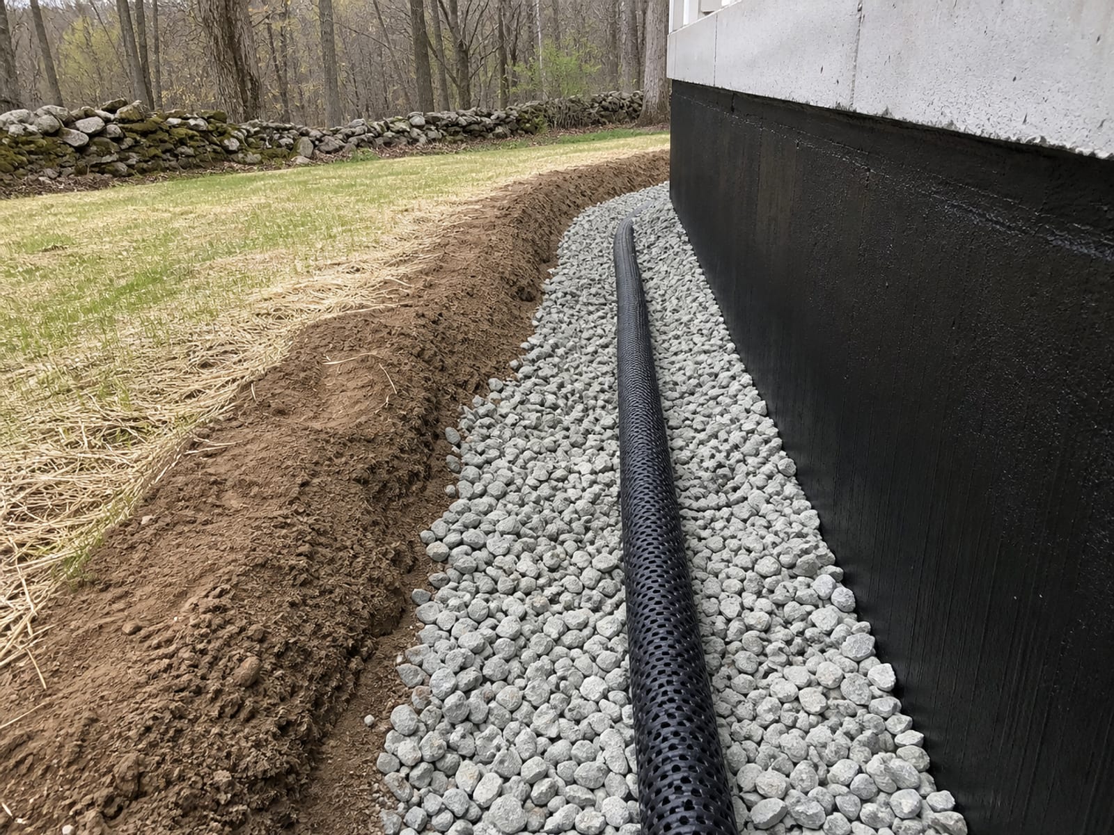 Perforated footing drain bedded in stone against a poured foundation wall with dampproofing applied, ready for inspection before backfill