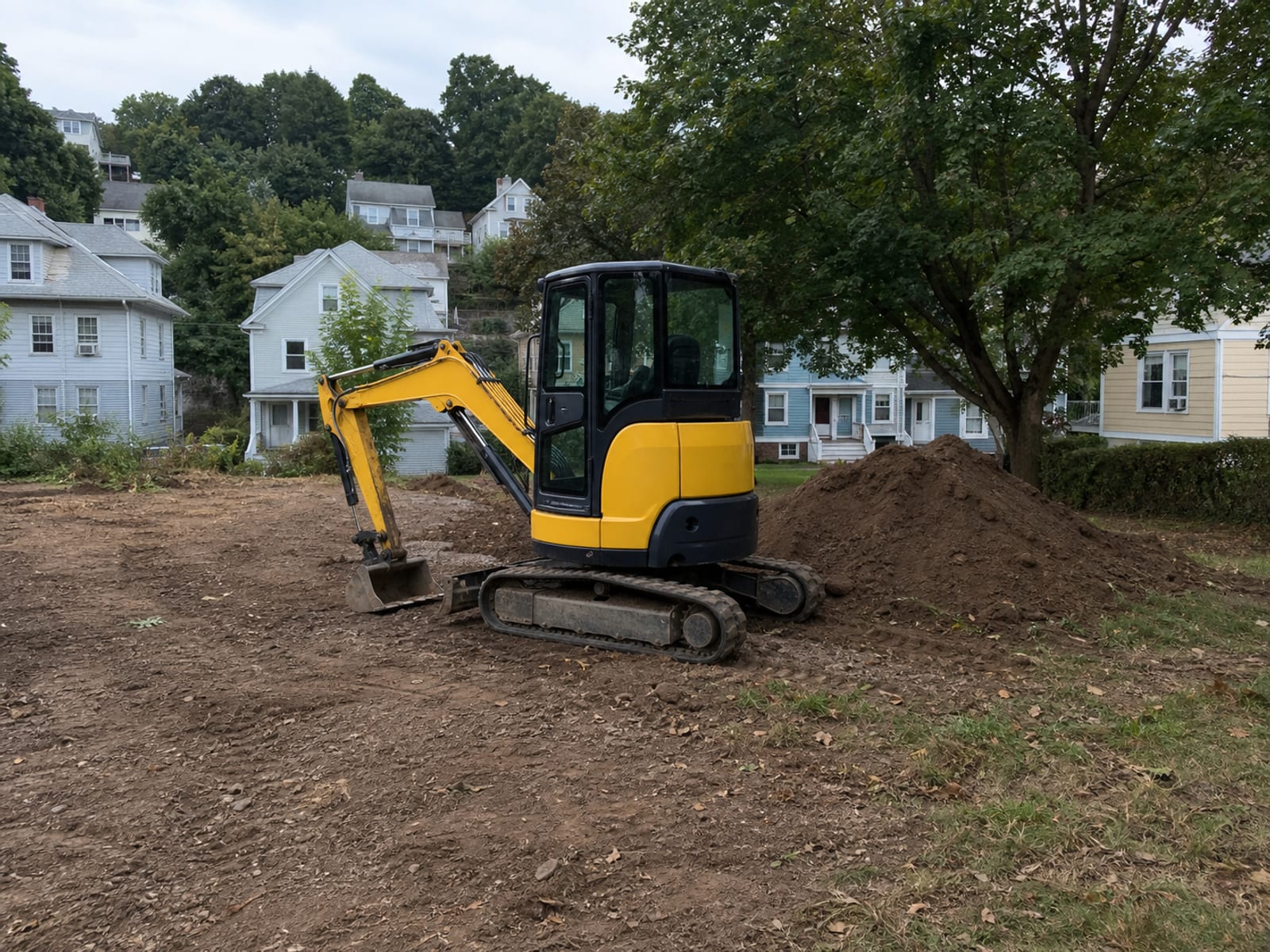 Tracked excavator parked at the edge of a rough graded building pad on a residential lot, bucket resting on the ground beside stockpiled topsoil