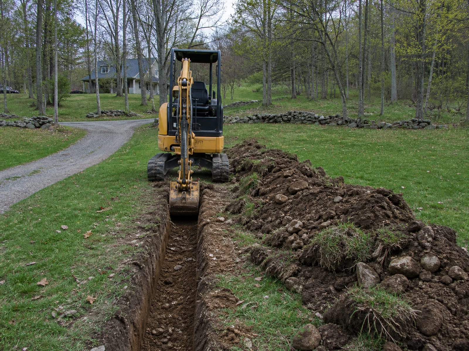 Compact excavator cutting a narrow utility trench across a rural property, with excavated soil windrowed well back from the open edge