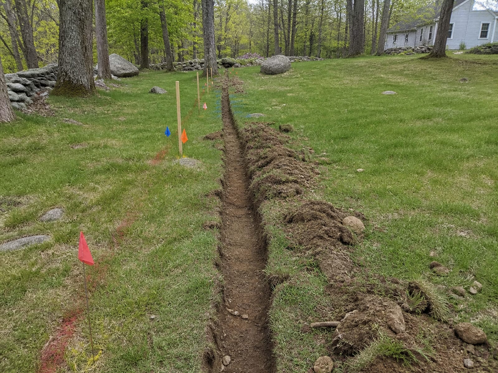 Utility locate paint marks and marker flags running across grass along a planned trench line, with staked route markers set alongside