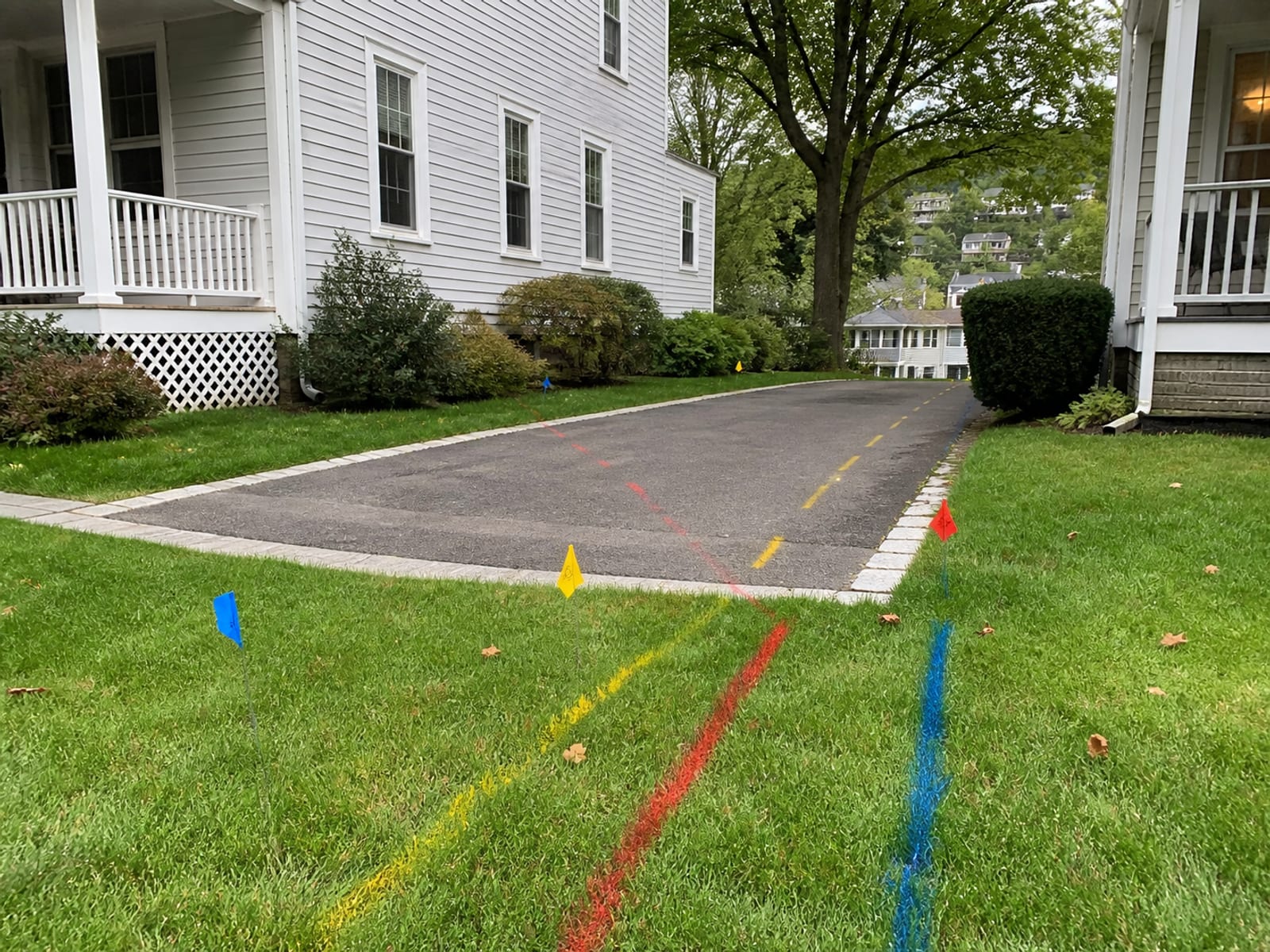 Colored utility locate paint lines and marker flags running across a residential lawn and driveway apron ahead of a trenching job