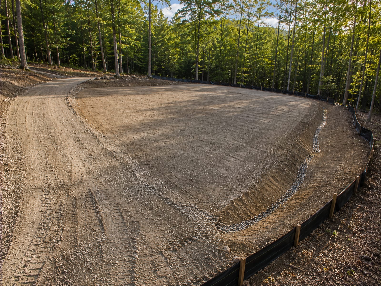 Finished graded and compacted building pad with rough graded driveway approach, drainage swale, and cleared wooded lot perimeter on a Connecticut residential property ready for foundation forming