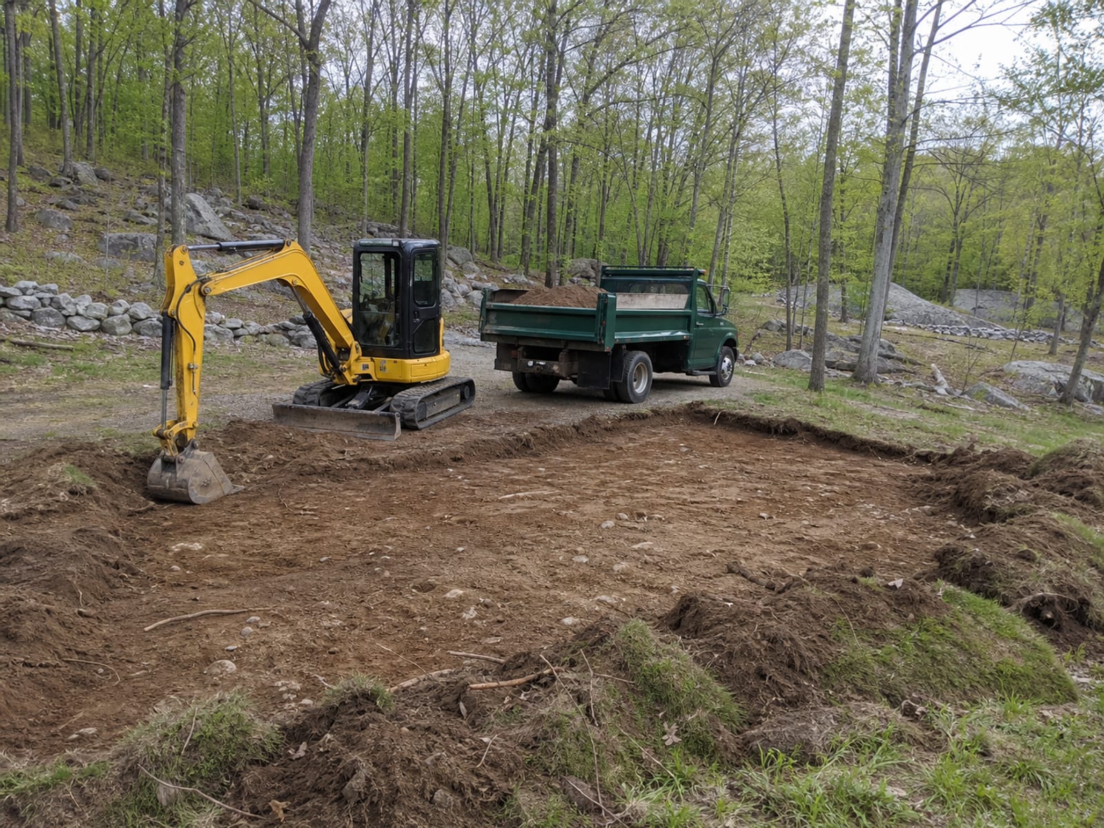Tracked excavator parked and idle beside an open excavation on a wooded rural parcel, bucket resting on the ground, a dump truck standing alongside part loaded with spoil