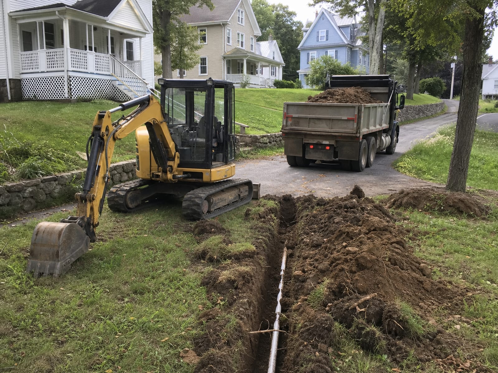 Compact excavator parked and idle in an open side yard beside a shallow excavation, bucket resting on the ground, a truck standing on the driveway part loaded with spoil