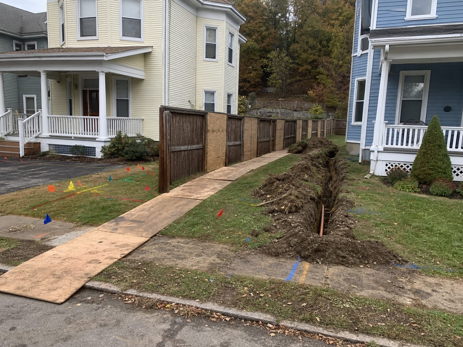 Colored utility locate marks across a lawn and driveway apron with plywood laid over the access route and fencing protected with boards