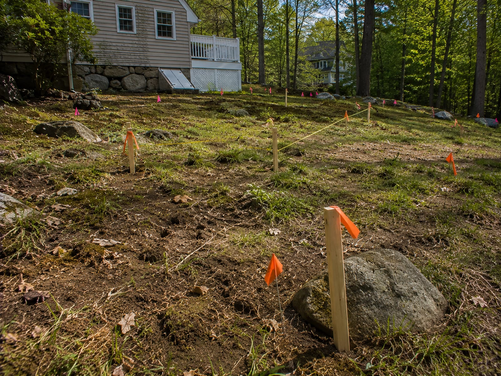 Residential yard with orange and pink survey flags and wooden stakes marking planned excavation boundaries on uneven terrain