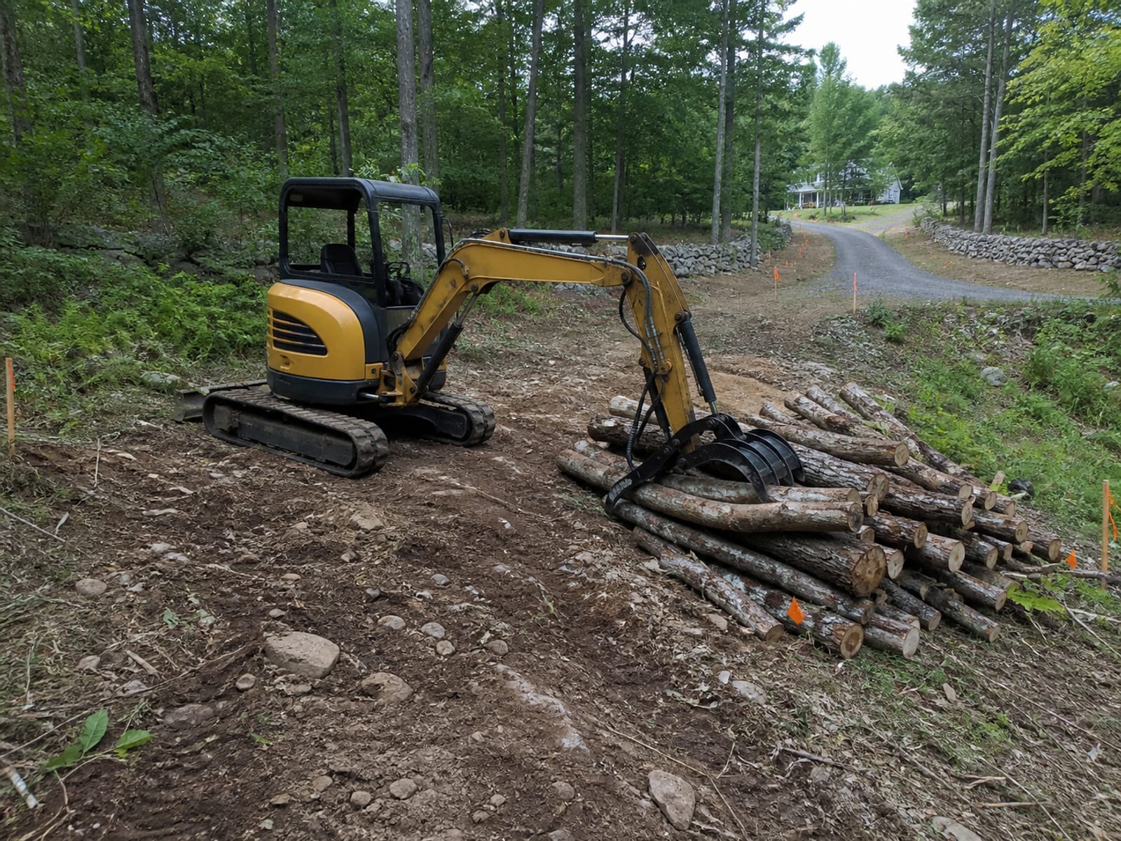 Tracked excavator with a grapple attachment lifting cut trunks onto a sorted pile on uneven wooded ground inside a flagged work area