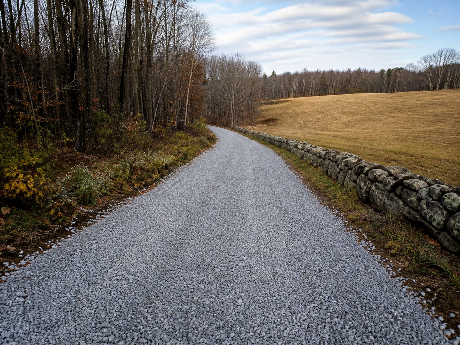 Rural private gravel drive rebuilt with fresh crushed stone running out of woodland past a dry stacked stone wall toward an open field