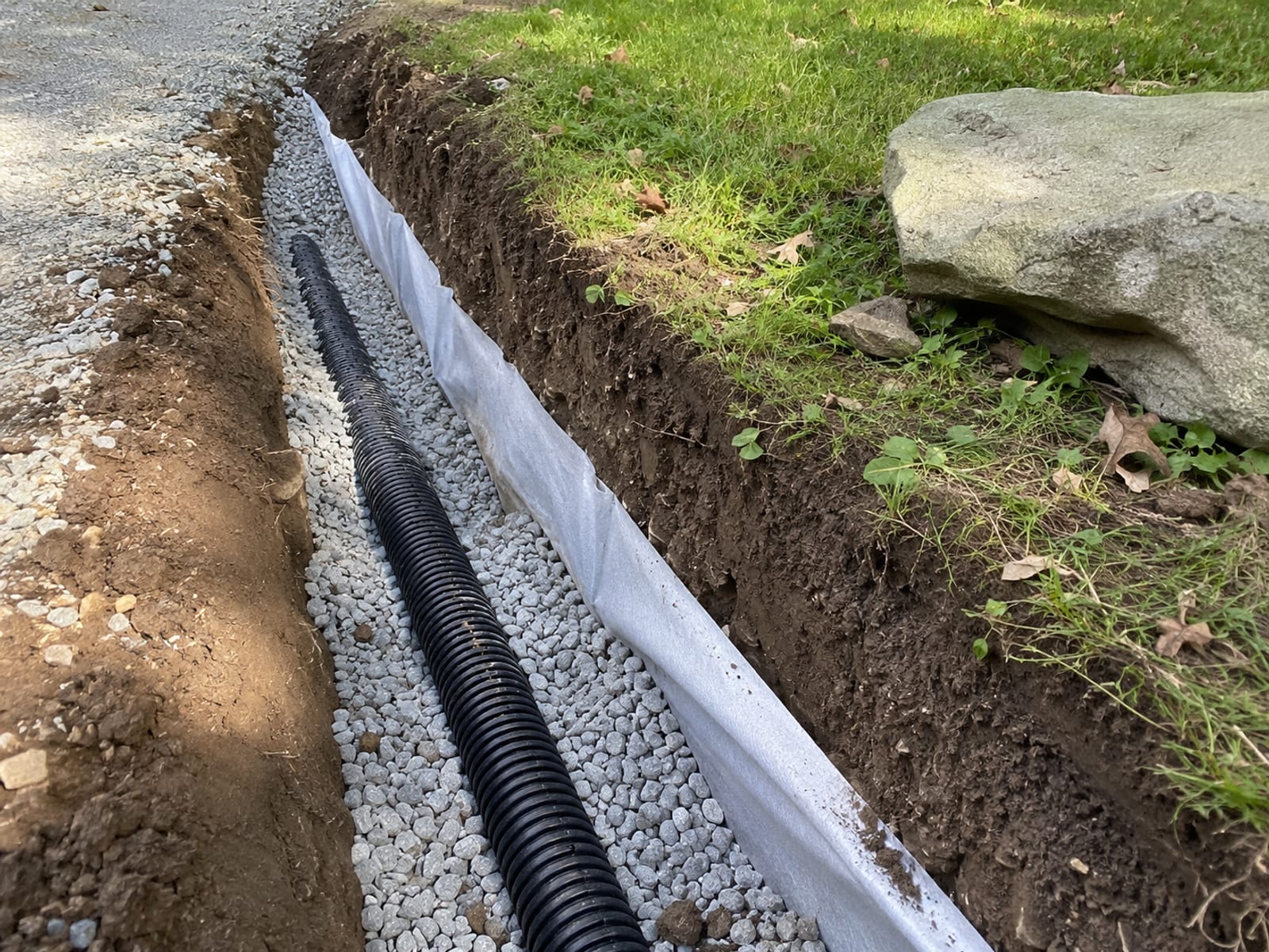 Freshly installed French drain trench with perforated pipe and washed gravel alongside a residential driveway in Connecticut