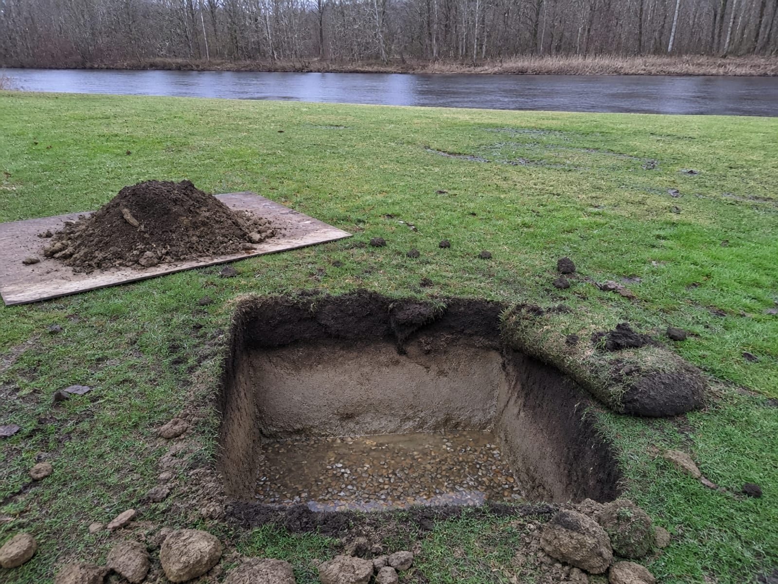 Small shallow test cut in a low lying riverside lawn showing damp silt over gravel with a little water collected in the bottom