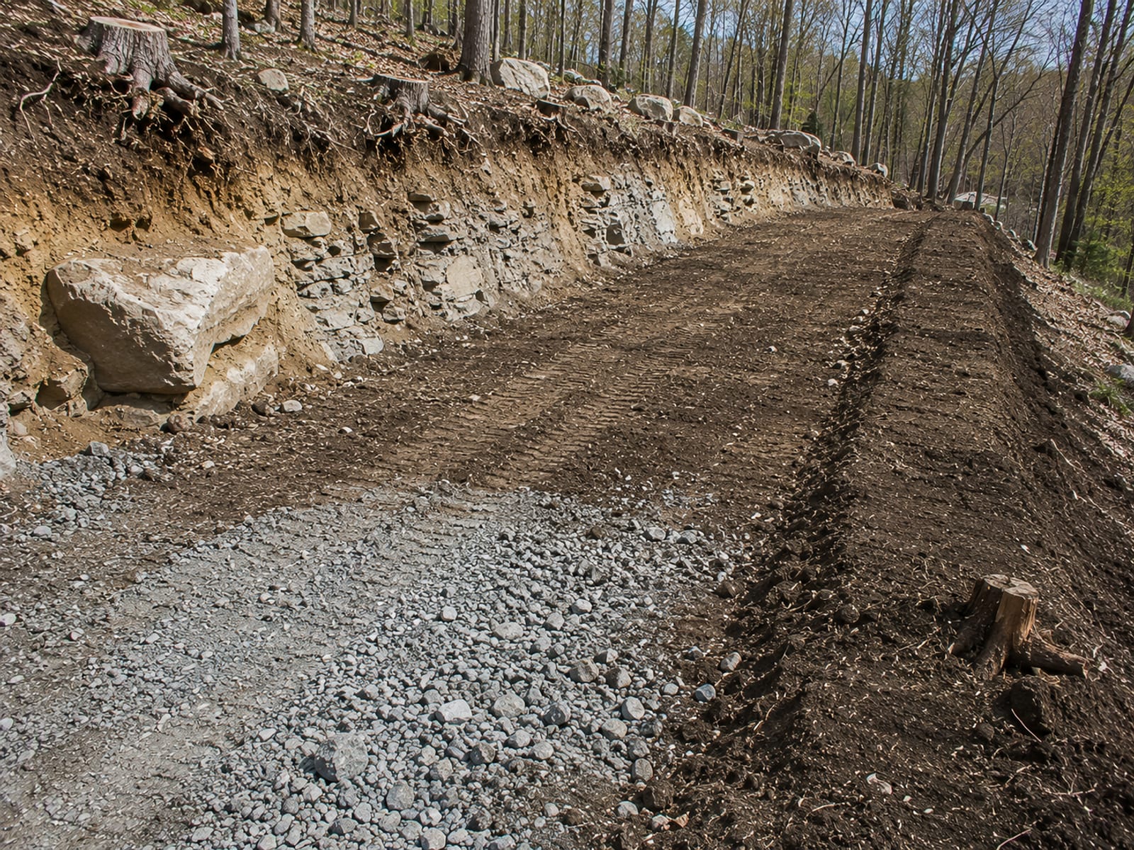 Excavated roadway subgrade on a sloped Connecticut lot showing cut and fill earthwork with exposed ledge rock and compacted soil berm