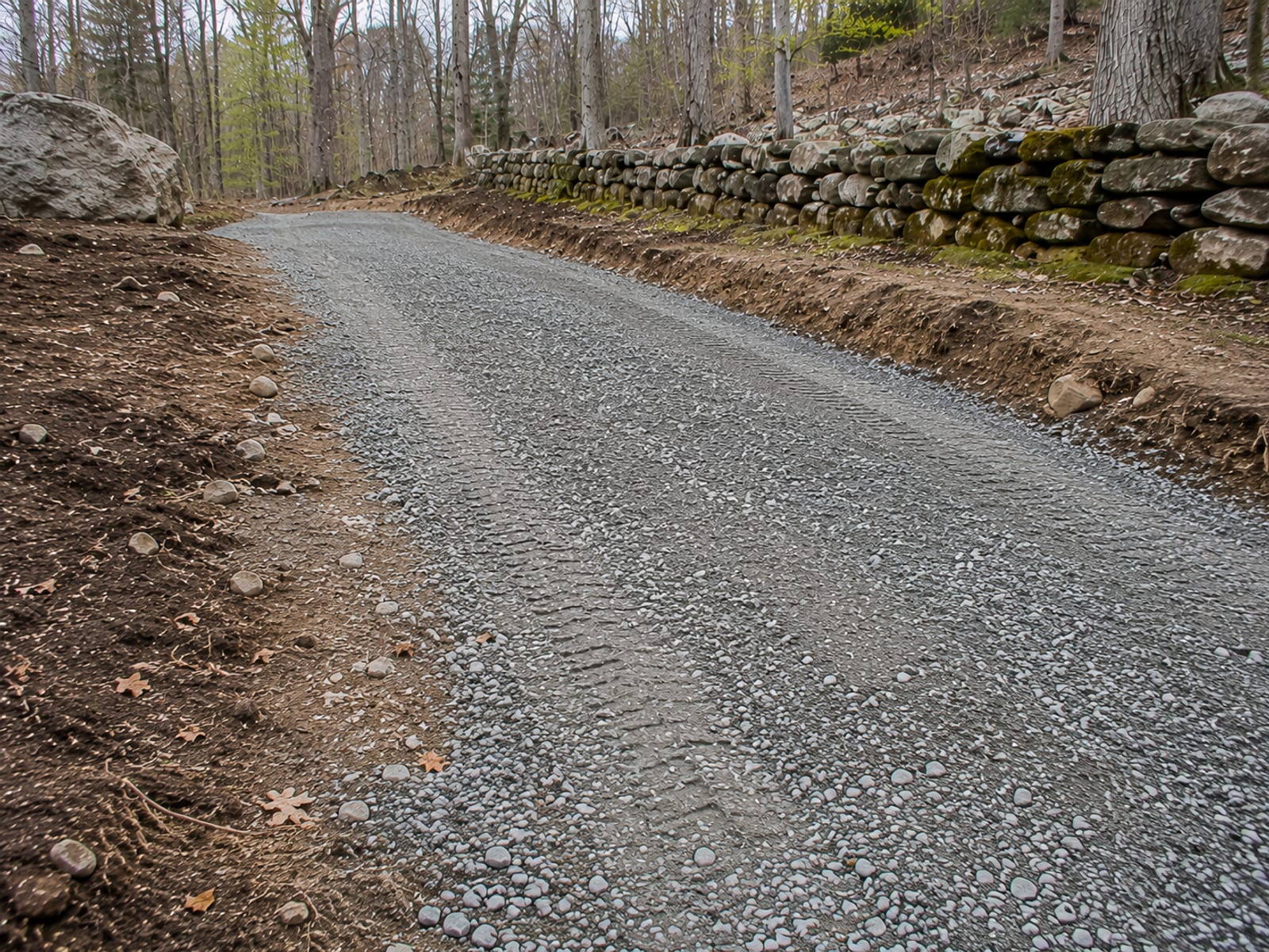 Freshly excavated driveway trench cut into sloped wooded lot with exposed glacial till soil and gravel subbase visible