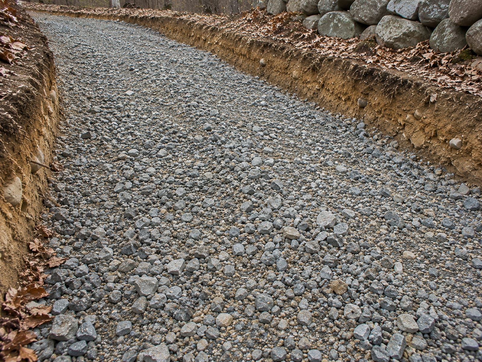 Close view of crushed stone and processed gravel subbase material spread across an excavated driveway bed with earthen cut walls on both sides