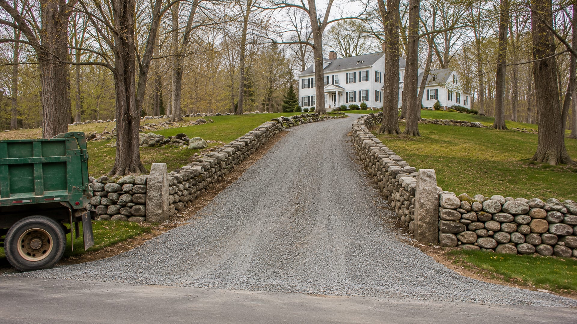 Wide view of a freshly excavated and gravel-surfaced driveway leading to a colonial style New England home with stone walls and deciduous trees
