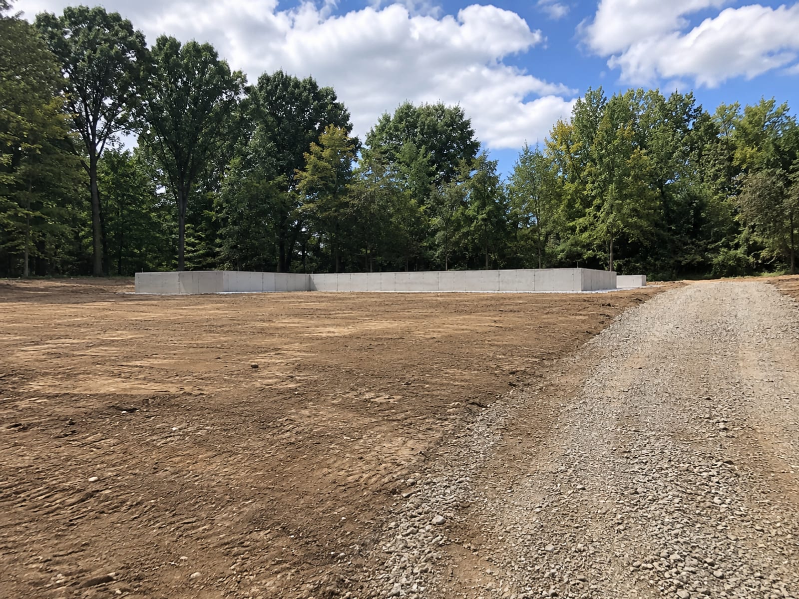 Fully excavated and cleared residential lot with smooth graded earth, a concrete foundation edge visible, and wooded backdrop in Connecticut