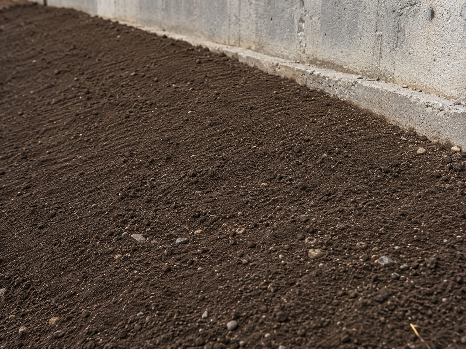 Close-up of freshly graded dark topsoil sloping away from a home foundation with embedded small stones