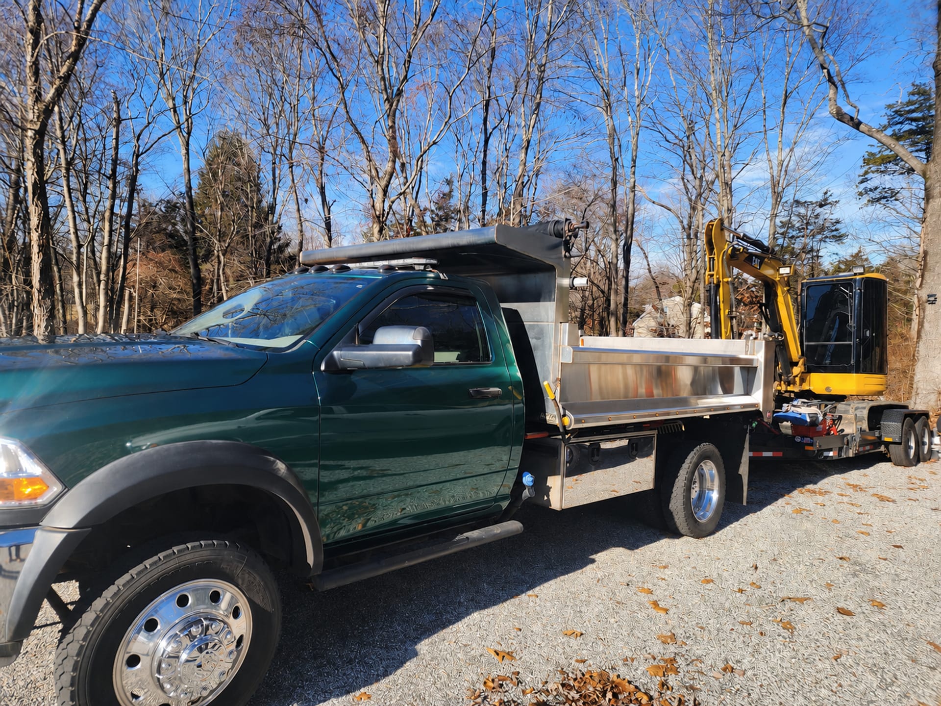 Green dump truck towing a yellow tracked excavator on a gravel lot with bare winter trees and blue sky