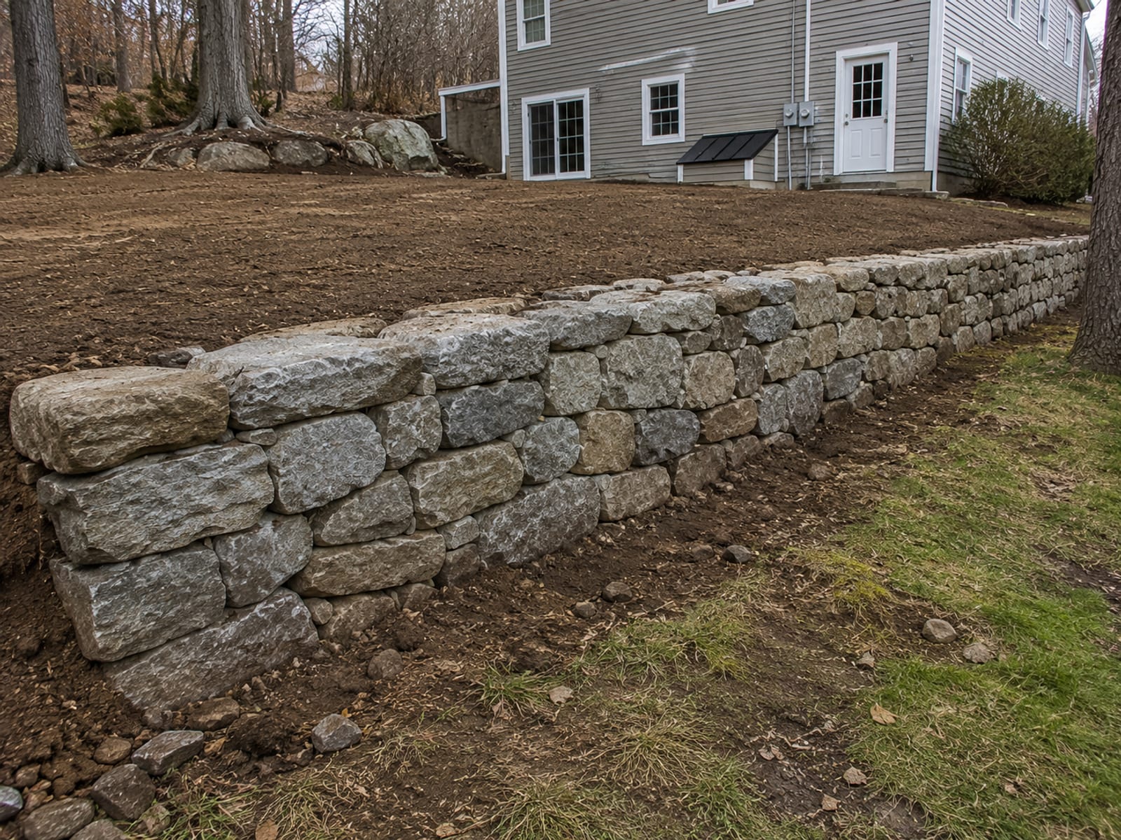 Newly constructed dry-stacked stone retaining wall holding back a terraced hillside slope on a residential lot in Connecticut