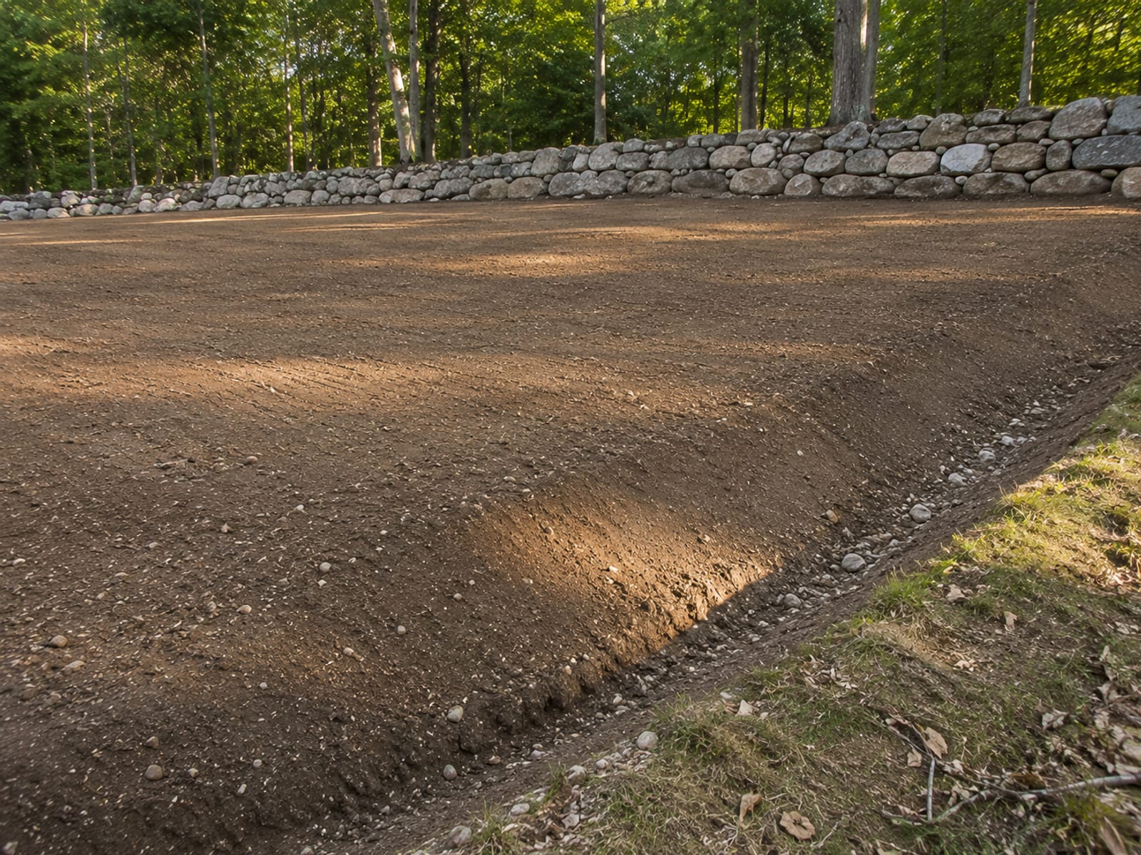 Freshly leveled and graded residential lot surface showing smooth compacted soil with a graded swale channel and stone wall at the property edge