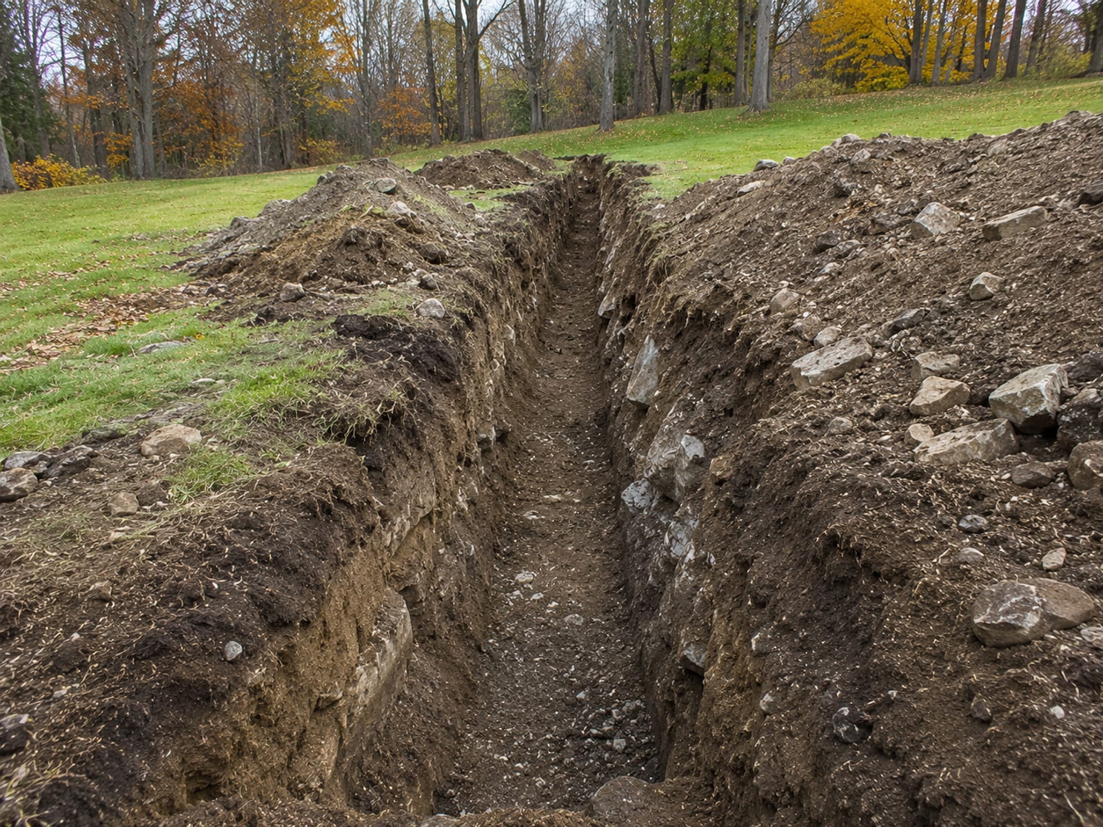 Freshly excavated utility trench cut through a rocky residential backyard in Connecticut