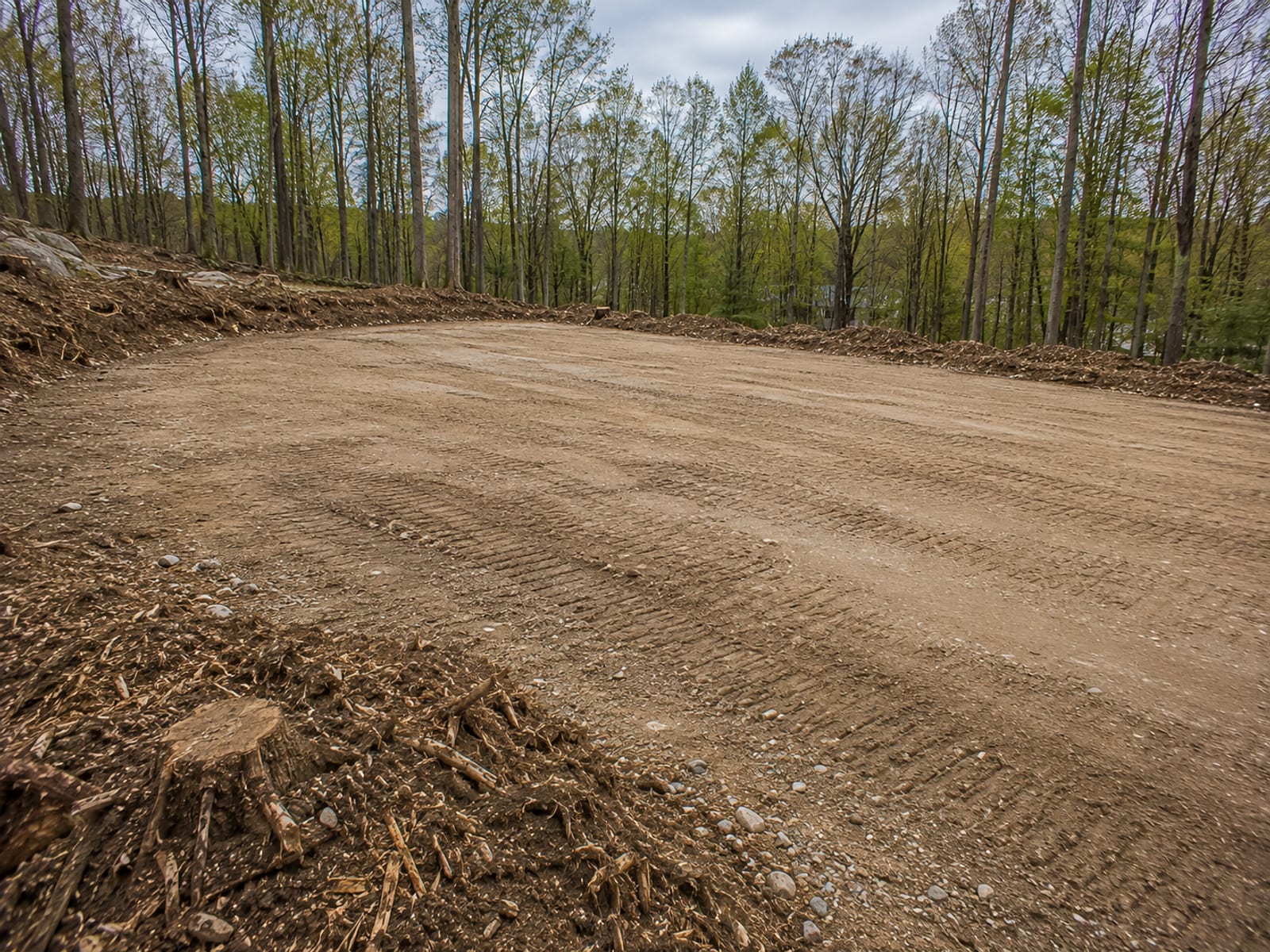 Freshly cleared and rough-graded level building pad cut into a formerly wooded sloped lot in Watertown CT with stumps removed and piles of mulched brush at the perimeter